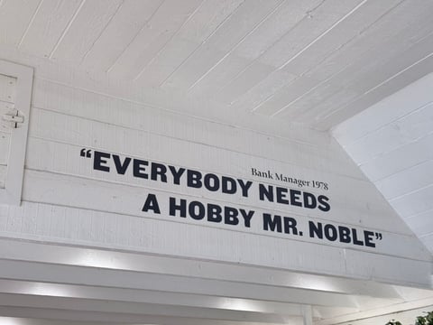 Sign on white ceiling displaying a quote: Everybody needs a hobby Mr. Noble, attributed to Bank Manager 1978