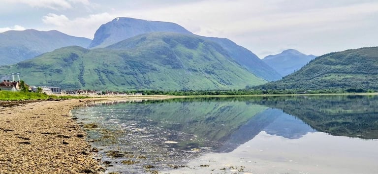Calm lake reflecting snow-capped mountains with green slopes, pebble beach in foreground, and small settlement on hillside