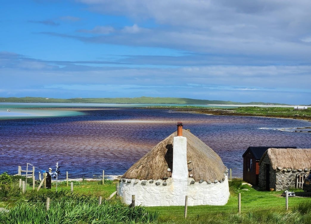 Traditional thatched-roof boat house with white trim beside a calm water body in a grassy Scottish landscape