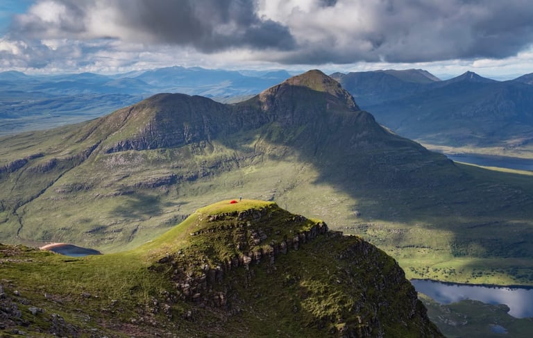 Aerial view of mountain peaks with green valleys, moorland, and lakes under cloudy sky in Scottish Highlands