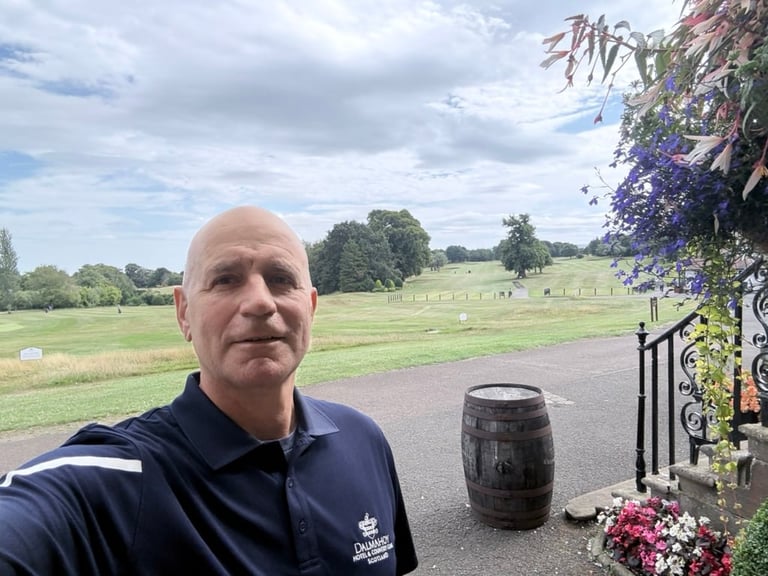 Bald man in navy polo shirt smiling at camera with rural pasture landscape and purple flowering vines in background