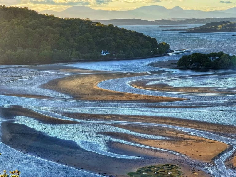 Scenic coastal landscape with sandy tidal flats, forested headlands, and mountains at sunset