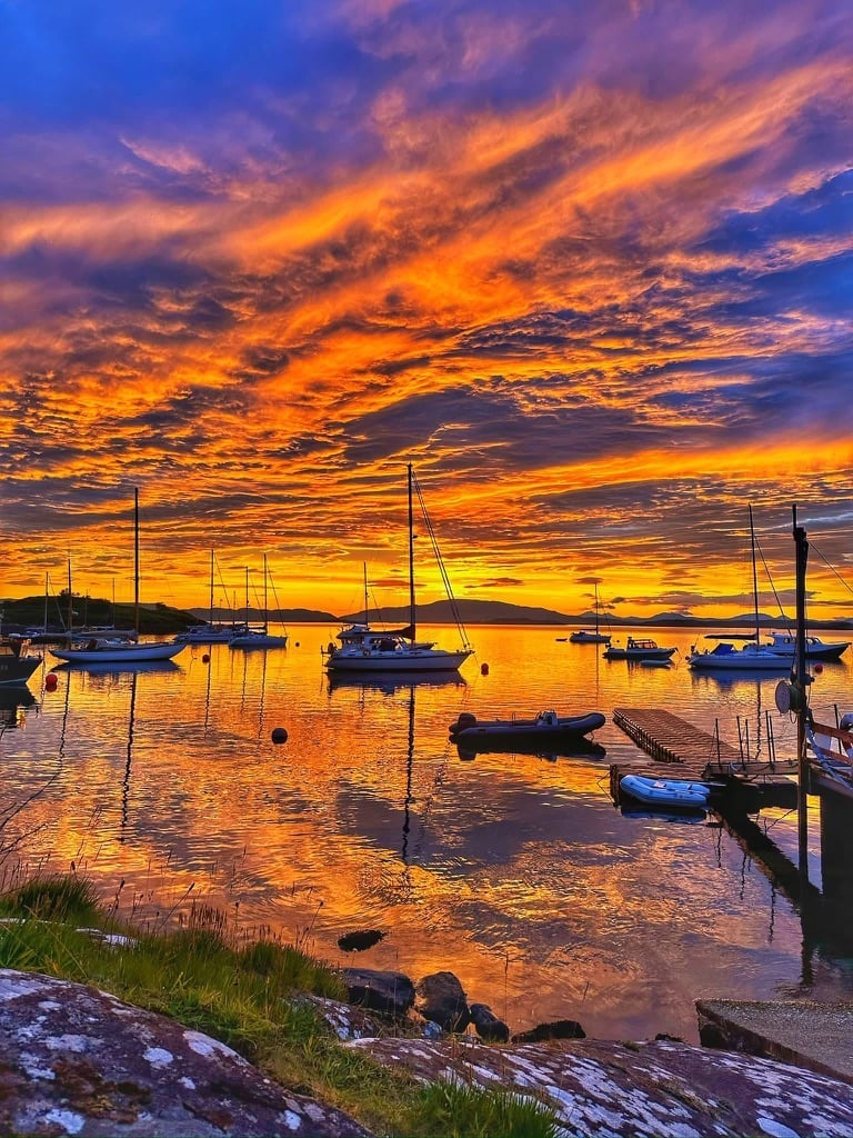 Sailboats moored in calm water at golden sunset with vibrant orange and blue sky reflected on the bay