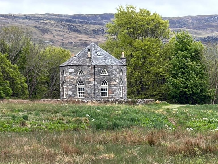 Abandoned stone chapel with Gothic-style windows surrounded by green fields and trees with hills in the background