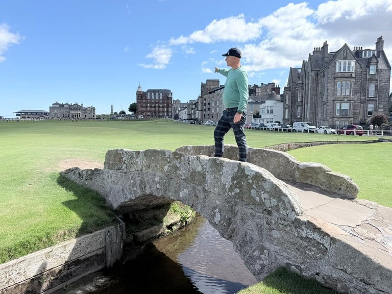 Person in green shirt standing on historic stone bridge over water with golf course and Scottish buildings in background