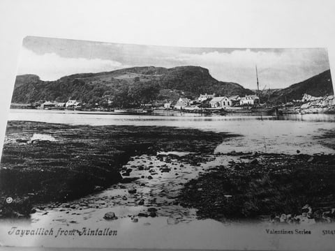 Black and white vintage photograph of a coastal village nestled beneath forested hills, with boats in the harbor and rocky shore in foreground
