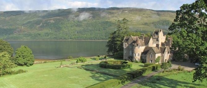 Ruins of a Scottish castle on a green hillside overlooking a loch, surrounded by trees