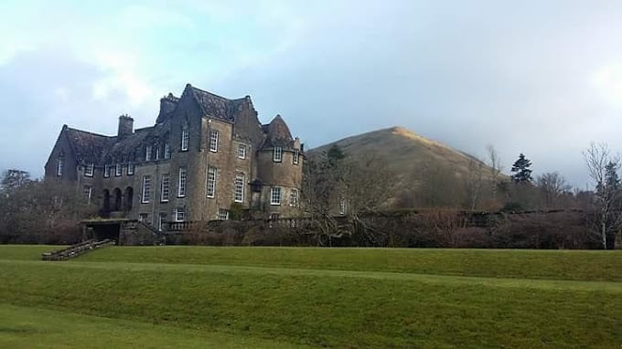 Large stone castle on sprawling green lawn with mountains in background under cloudy sky