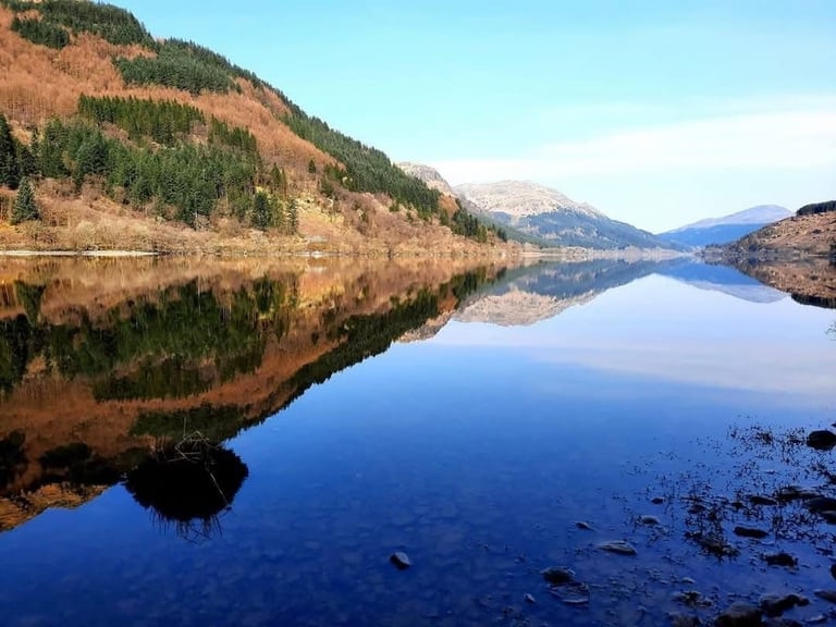 Calm alpine lake with mirror-like reflections of forested mountains and snowy peaks under clear blue sky