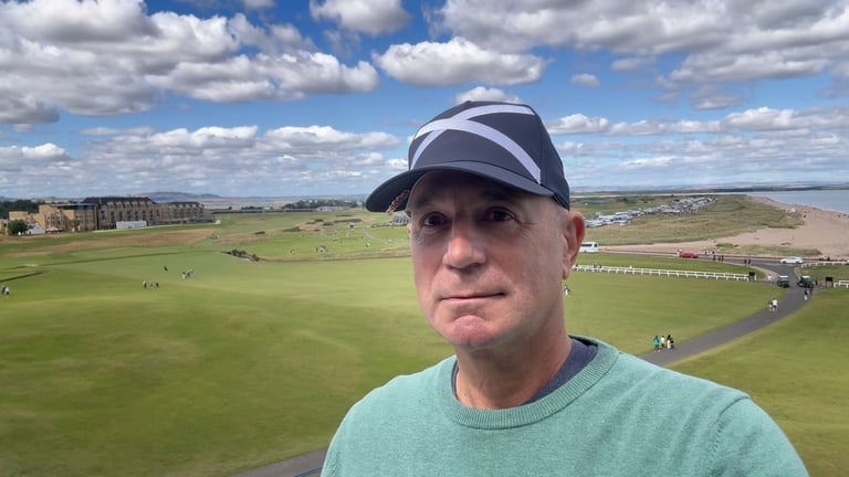 Man wearing a blue and white baseball cap and light blue shirt smiling at a golf course with green fairways and blue sky with clouds in background