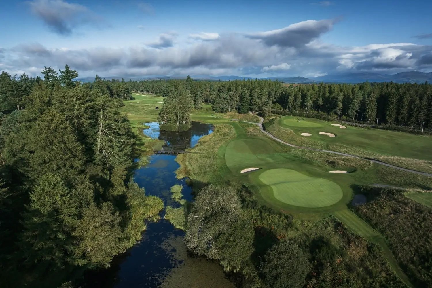 Aerial view of a golf course with manicured greens, fairways, water hazards, and dense coniferous forest under a cloudy sky