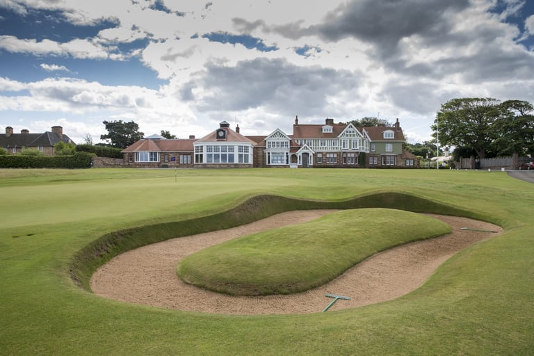 Championship golf course with large sand bunker in foreground and historic clubhouse building across the fairway under blue cloudy sky
