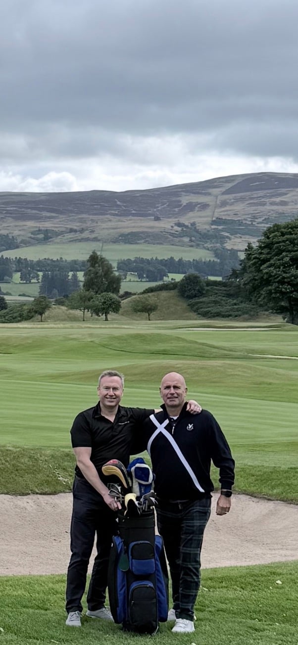 Two men stand at a golf course with a golf bag, smiling at the camera with rolling hills and green landscape in the background