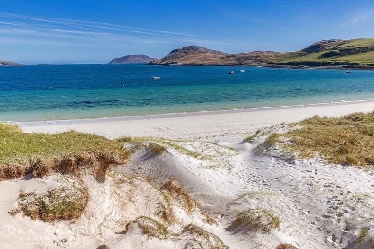 Scenic beach with white sand, turquoise water, and rolling mountains under a clear blue sky