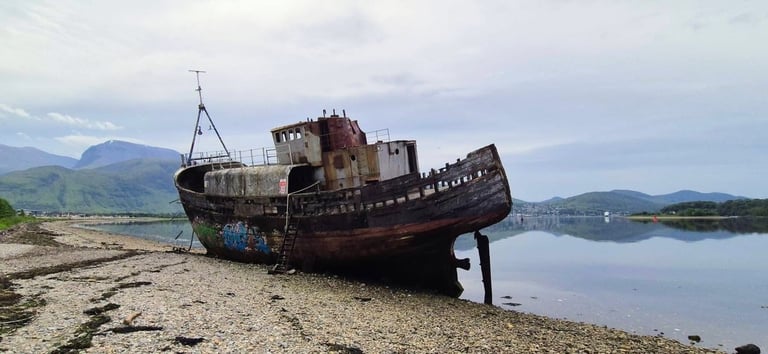 Abandoned rusty shipwreck beached on rocky shore with mountains and water in background