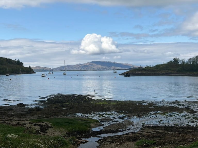 Scenic coastal landscape with calm blue water, sailboats anchored in a bay, forested shorelines, and mountains under a cloudy sky