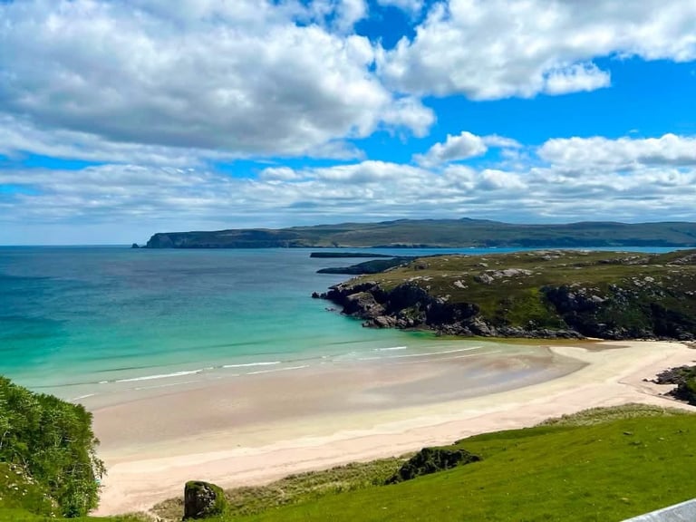 Scenic coastal landscape with sandy beach, turquoise waters, green cliffs, and islands under blue cloudy sky