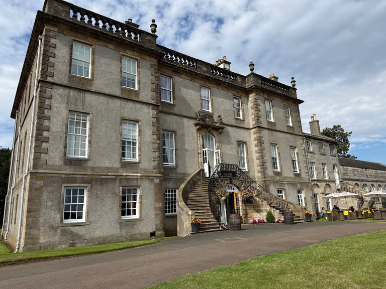 Grand stone manor house with multiple windows, exterior staircase, and manicured grounds under cloudy sky