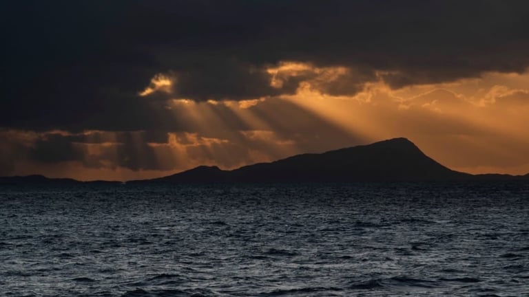 Dramatic sunset over dark ocean with silhouetted volcanic island and golden clouds breaking through stormy sky