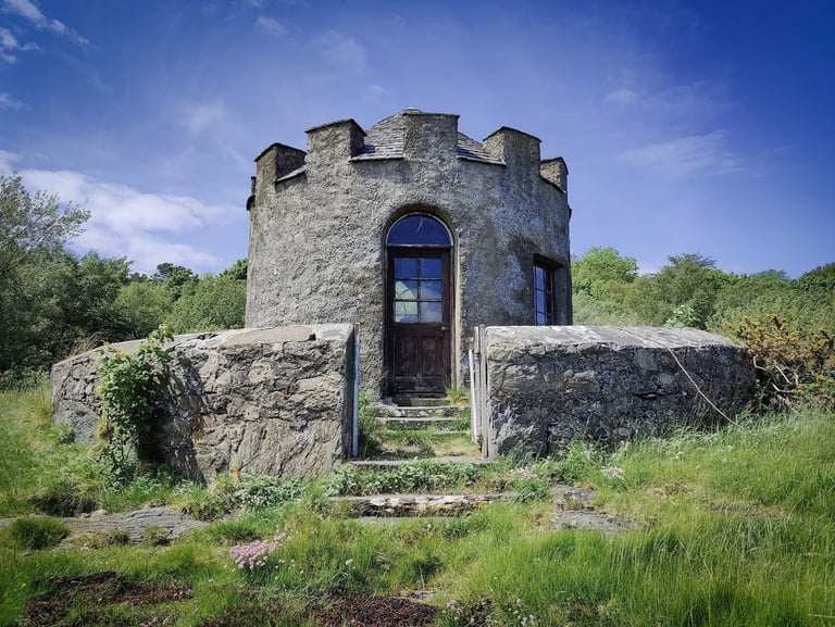 Historic stone tower with crenellated top and blue door, surrounded by grass and green vegetation under blue sky