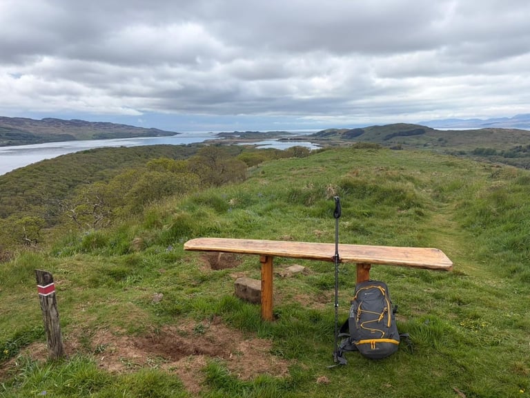 Wooden bench on hillside overlooking mountains and lakes under cloudy sky, with hiking gear nearby