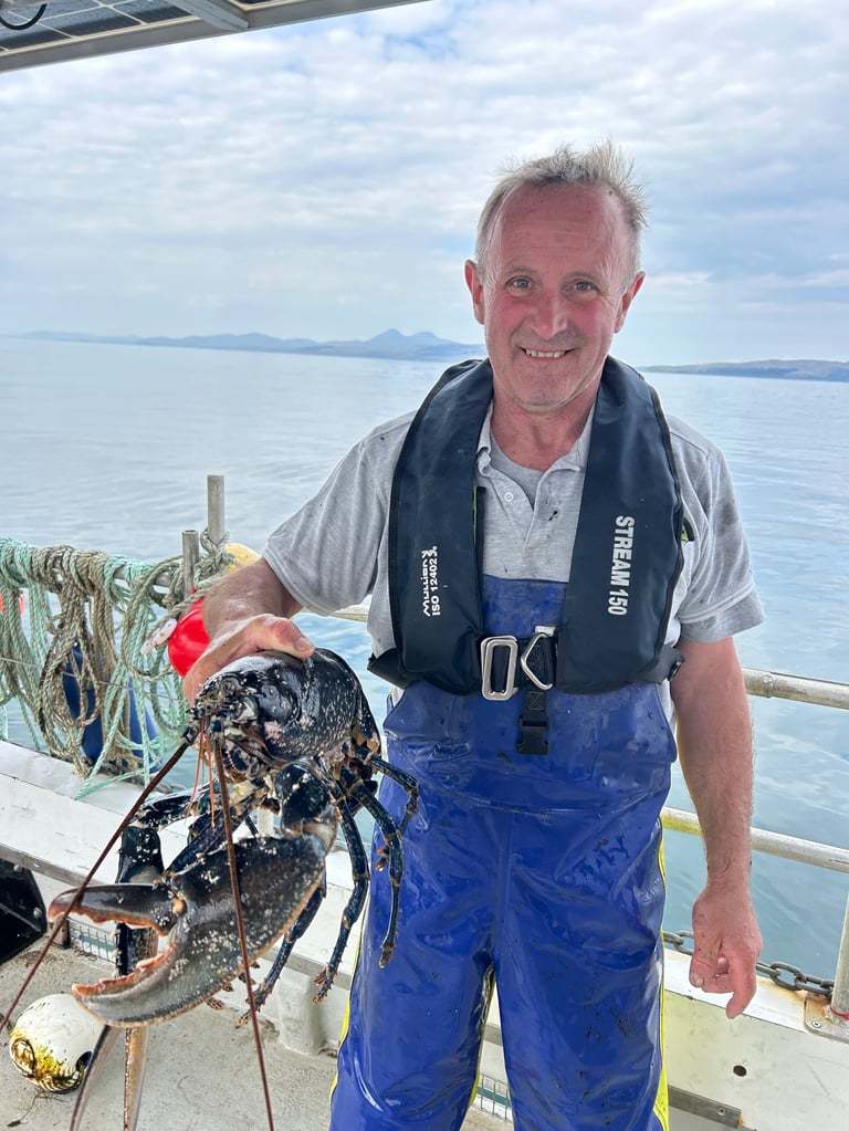 Man in blue overalls holding a large lobster on a boat with calm ocean and mountains in the background