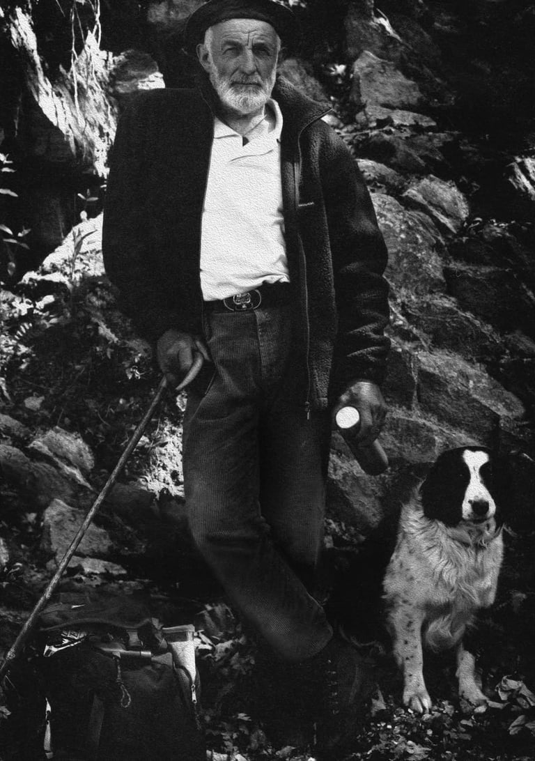 Elderly man in dark coat and cap standing with a small dog among rocky terrain in black and white photograph