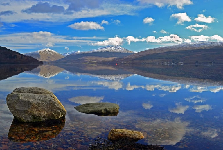 Still mountain lake with perfect sky reflection, boulders in shallow water, and snow-capped peaks in distance