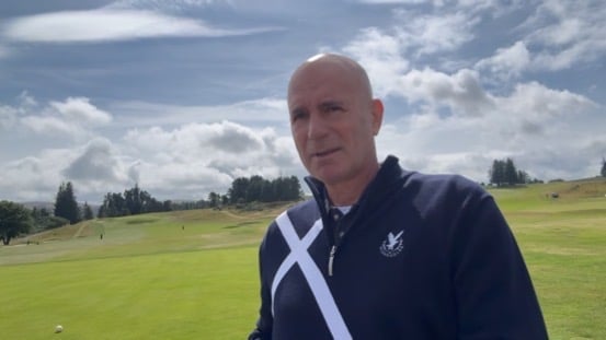 Bald man in dark blue golf jacket with white X logo on a scenic golf course with green fairways and mountains in the background