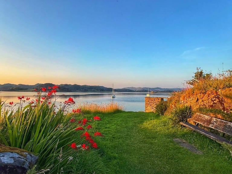 Red flowers blooming on a grassy lakeside garden overlooking calm blue water with sailboat and mountains at sunset