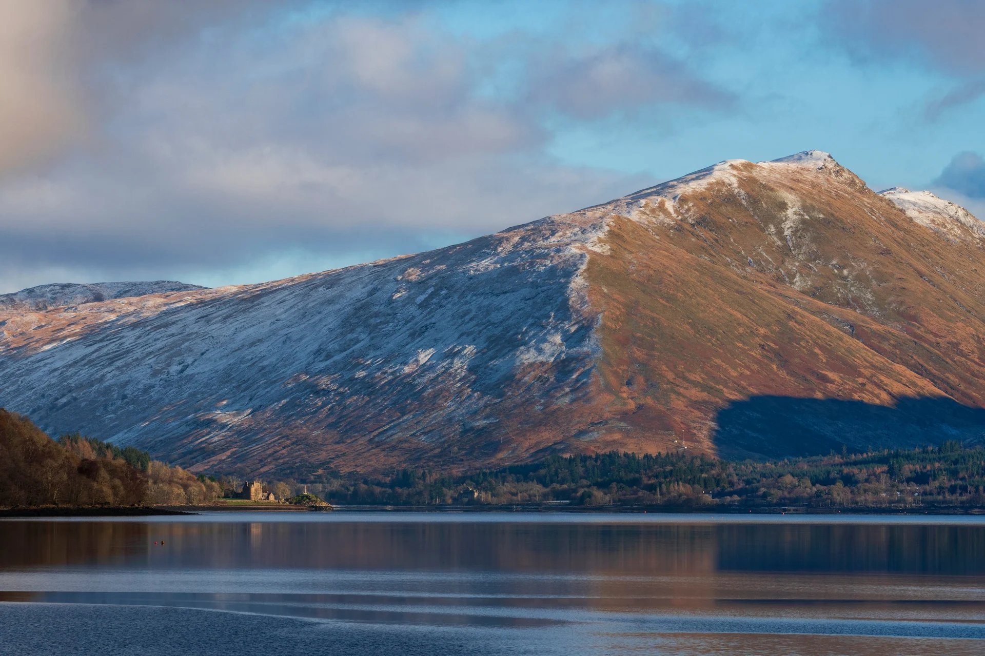 Snow-capped mountain reflected in calm lake with forested shoreline beneath blue sky
