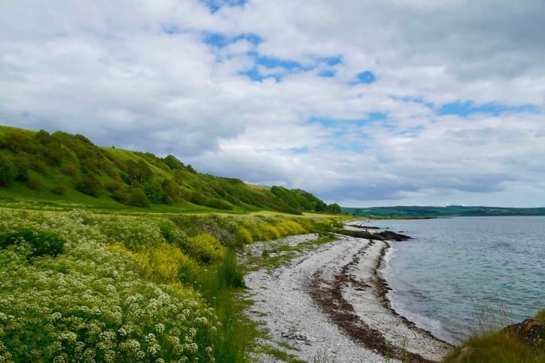 Coastal landscape with green grassy cliffs, pebble beach, and calm blue water under a partly cloudy sky