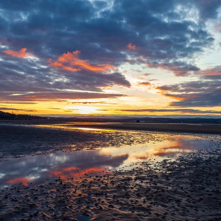 Dramatic sunset over a beach with golden and orange sky reflected in wet sand and shallow water