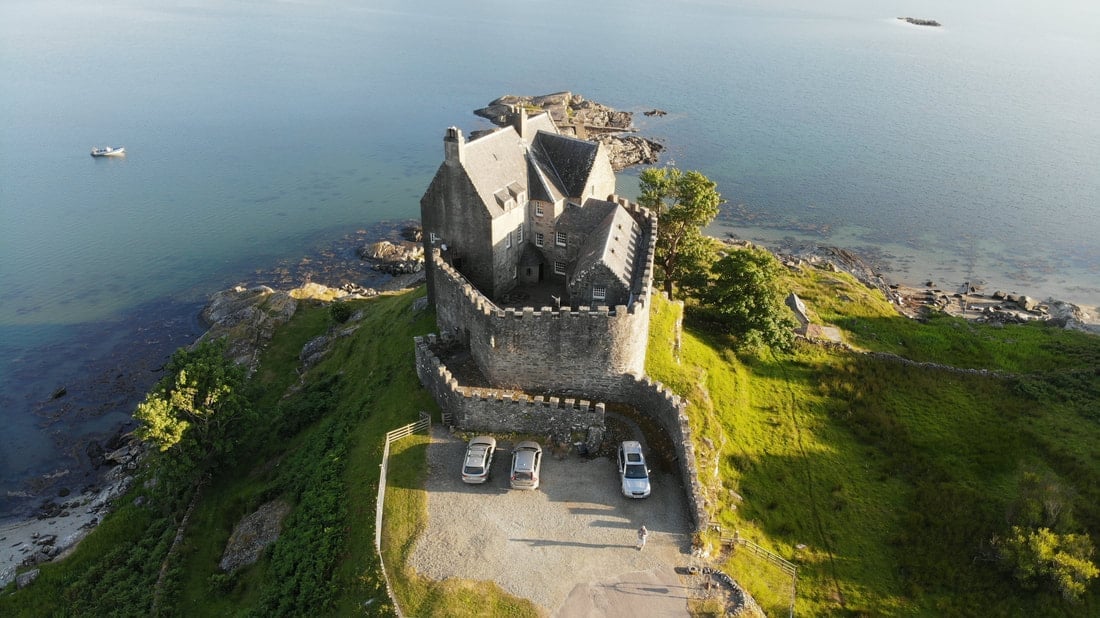Aerial view of an ancient stone tower on a grassy cliff overlooking the sea with parked cars at its base