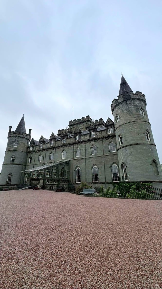 A grand Scottish stone castle with distinctive cylindrical towers and conical turrets, featuring a reddish gravel courtyard under a cloudy sky