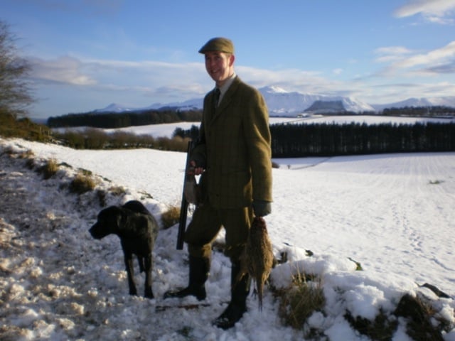 Man in olive jacket and cap standing with black dog in snowy mountain landscape with frozen lake