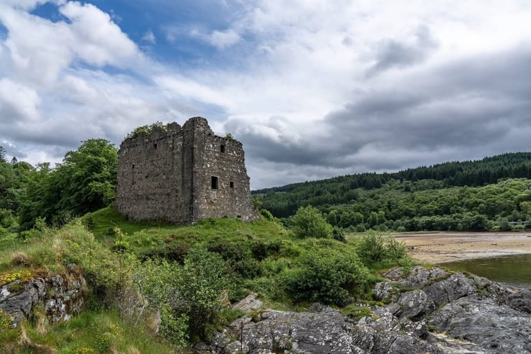 Ruined stone castle tower on rocky hillside overlooking a river valley with forested landscape and dramatic cloudy sky