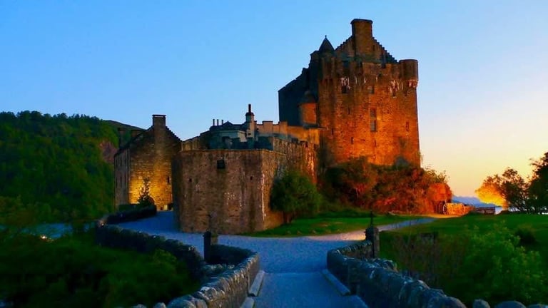 Illuminated medieval castle with stone towers reflected in moat at twilight, surrounded by green hills and trees