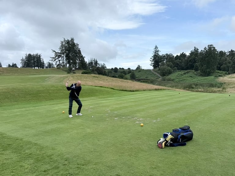Golfer in dark clothing mid-swing on a green fairway with golf bag nearby and scenic landscape with trees in the background