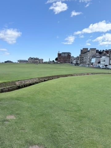 Historic golf course with manicured green turf and stone wall, lined with traditional buildings under blue sky