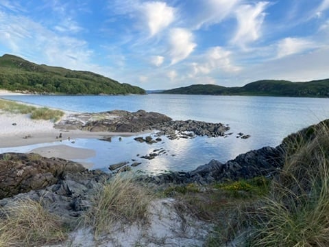 Scenic coastal beach with rocky outcrops, sandy shore, and forested hills under a blue sky with white clouds