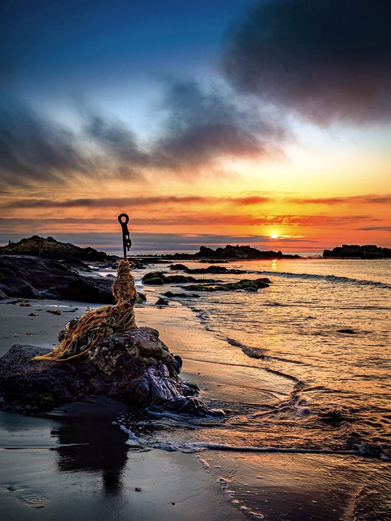 Person standing on rocky outcrop at beach during golden sunset with ocean waves and dramatic sky