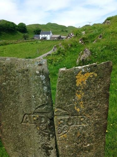Two weathered stone grave markers with lichen in a rural cemetery, with green hills and a white cottage visible in the background