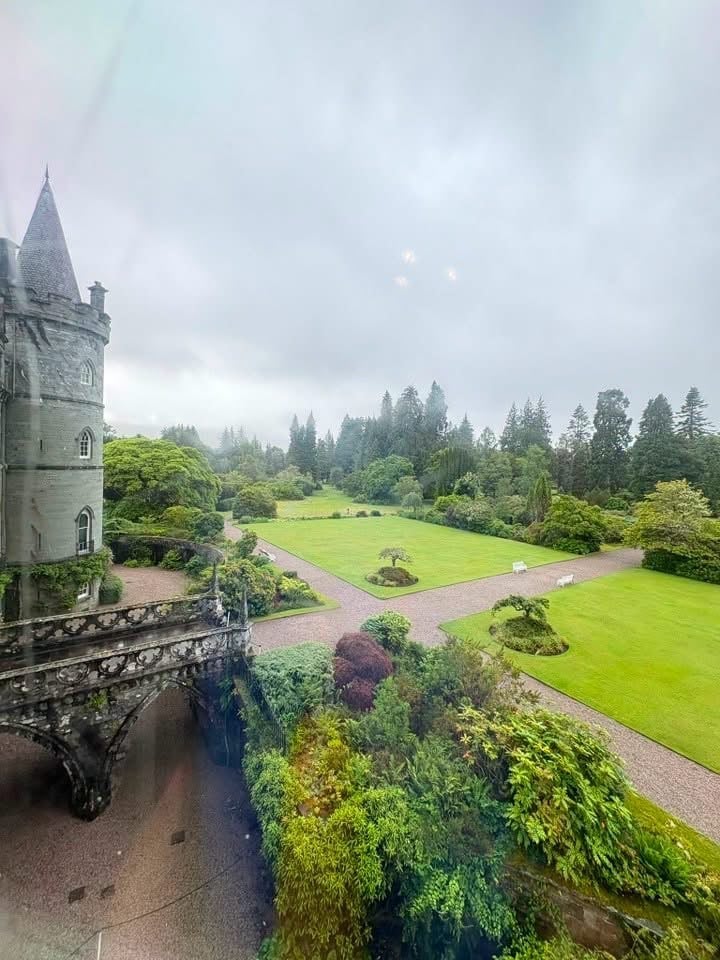Stone castle tower overlooking manicured grounds with green lawns, gardens, and evergreen trees under cloudy sky