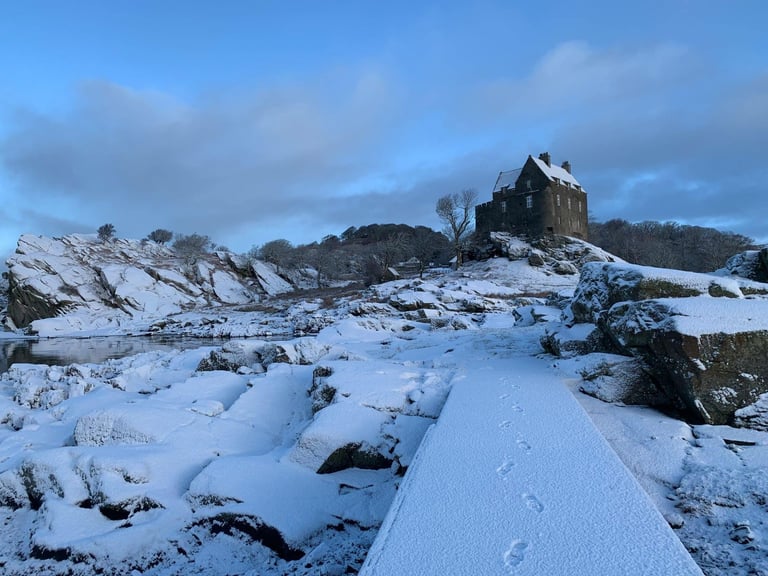 Snow-covered Scottish landscape with dark stone castle perched on a rocky hill under a blue winter sky