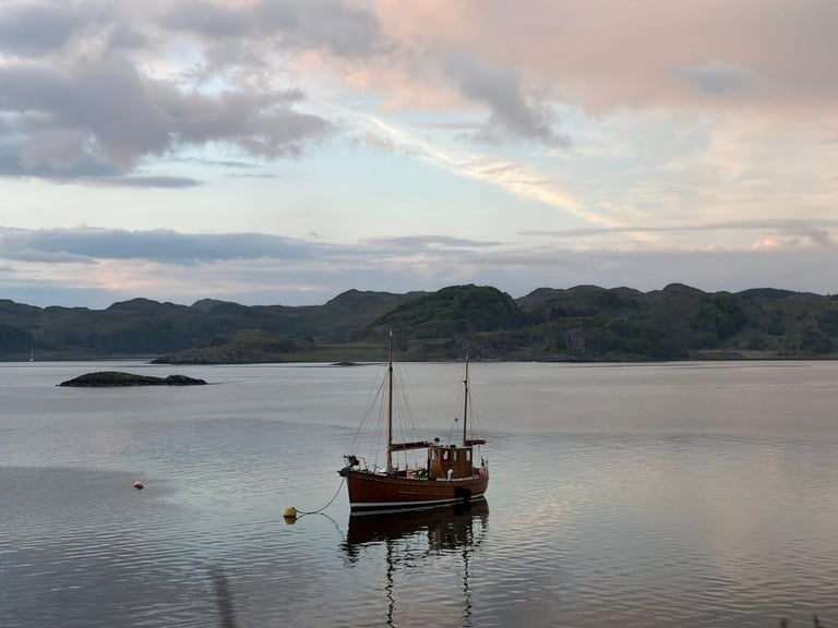 Red sailing boat moored in calm waters with green hills in the background under a cloudy sky