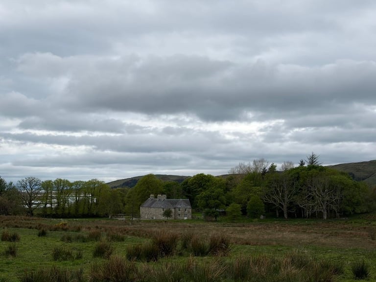 Abandoned stone cottage in a grassy Highland landscape with rolling hills and scattered trees under a cloudy sky