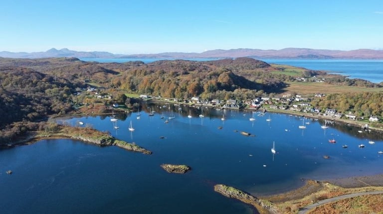 Aerial view of a coastal bay with sailboats anchored among rocky islands, surrounded by tree-covered hills and a distant mountainous landscape