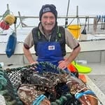 Person in blue apron holding up a large sea urchin on a boat dock with fishing equipment visible in background