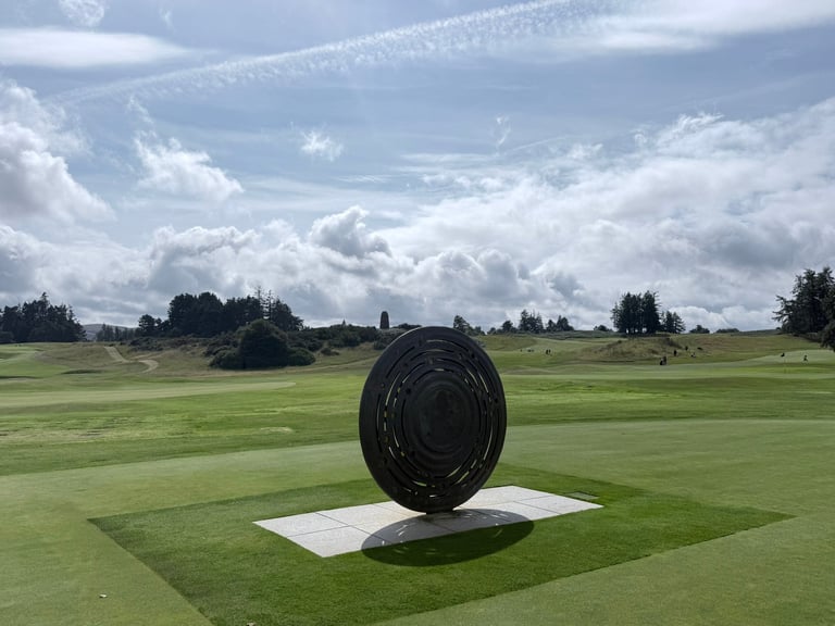 Large black spiral metal sculpture on white platform in green field under cloudy sky